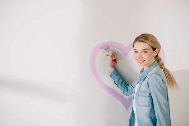 happy young woman painting pink heart with paintbrush on white wall - Photo, Image