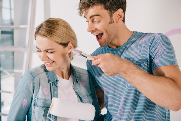 cheerful young man having fun with girlfriend while frightening with pink paintbrush - Photo, Image