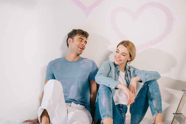 happy young couple sitting by white wall under drawn pink heart - Photo, Image