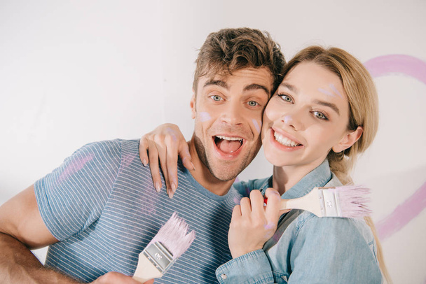 happy young couple hugging and smiling at camera while holding pink paintbrushes - Foto, immagini