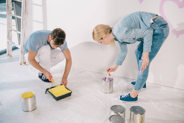 young man putting paint roller into yellow paint while girlfriend taking pink paint with paintbrush - Fotoğraf, Görsel
