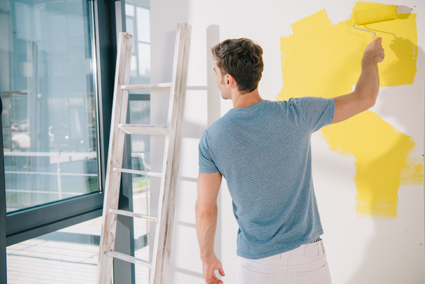back view of young man painting white wall in yellow with paint roller - Foto, afbeelding