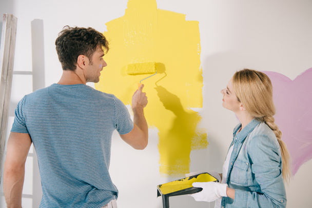 young woman holding roller tray while boyfriend painting wall in yellow with paint roller - Photo, Image