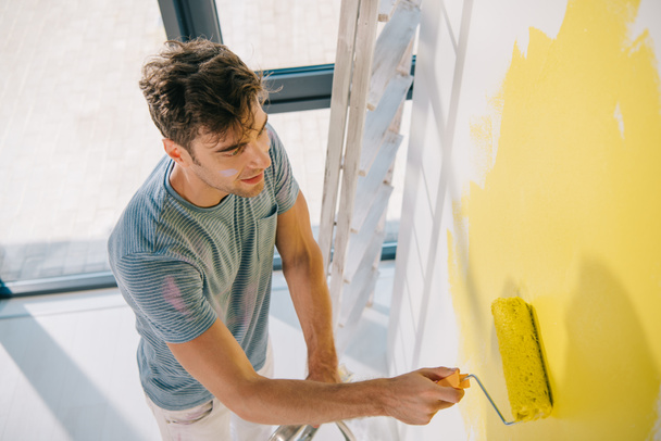 high angle view of handsome young man painting wall in yellow with paint roller - Valokuva, kuva