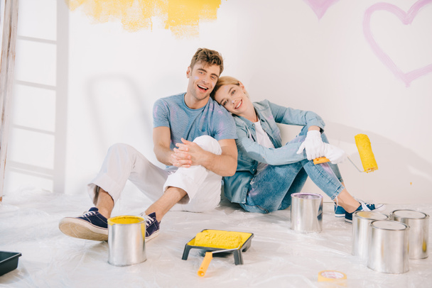 happy young woman holding yellow paint roller while sitting on floor near boyfriend - Photo, Image