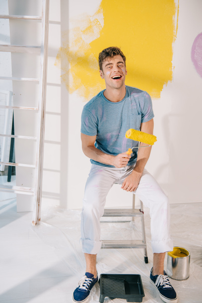 joven guapo sentado en la escalera, sosteniendo el rodillo de pintura amarillo y sonriendo a la cámara
 - Foto, imagen