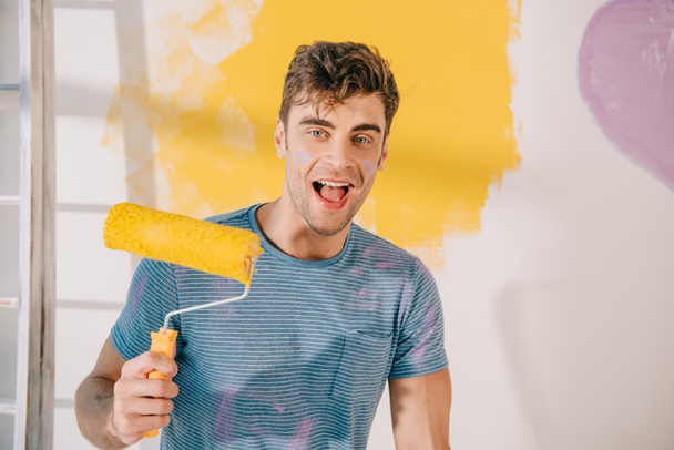 cheerful young man holding yellow paint roller while looking at camera - Photo, Image