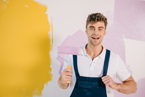 handsome young painter in overalls holding paint roller while standing near wall painted in yellow and pink - Fotografie, Obrázek