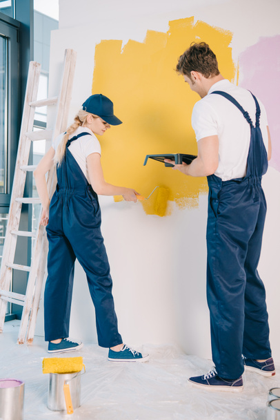 jonge schilder holding roller lade terwijl aantrekkelijke collega schilderen muur met verfrol - Foto, afbeelding
