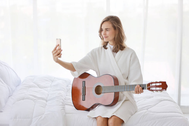 beyaz pijama genç bir beyaz kadın modern yatak odasında bir yatakta akustik gitar çalarken cep telefonundan kendini fotoğraf çekerken (teknoloji kavramı ile rahatlama) - Fotoğraf, Görsel