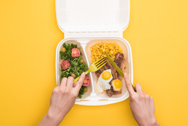 abgeschnittene Ansicht einer Frau, die aus einer Ökopackung mit Mais, Fleisch, Spiegeleiern und Salat isst, isoliert auf gelb  - Foto, Bild