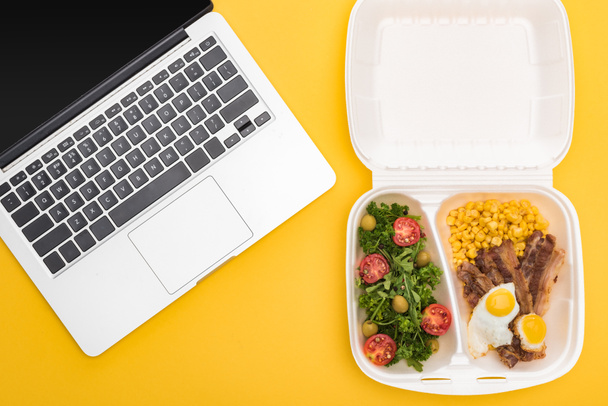 top view of laptop and eco package with corn, meat, fried eggs and salad isolated on yellow  - Photo, Image