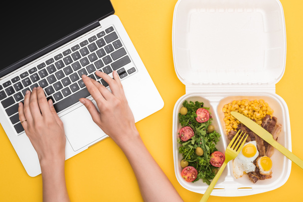 vista cortada da mulher usando laptop e pacote ecológico com milho, carne, ovos fritos e salada isolada em amarelo
  - Foto, Imagem