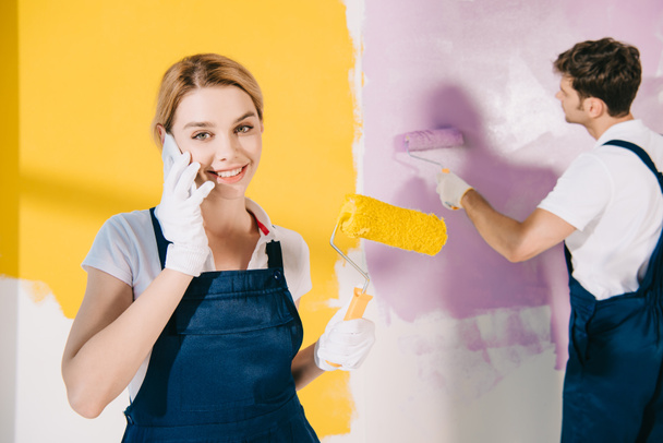 attractive painter in uniform talking on smartphone while handsome colleague painting wall - Фото, изображение