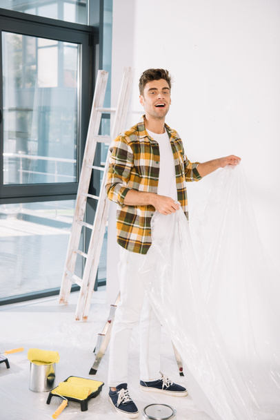 joven guapo sosteniendo celofán mientras se prepara para la pintura de la pared
 - Foto, Imagen