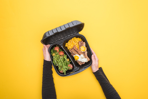vista cortada de mulher segurando pacote ecológico com milho, carne, ovos fritos e salada isolada em amarelo
  - Foto, Imagem