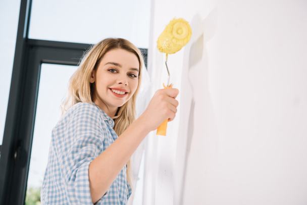 vrolijke jonge vrouw houden gele verf roller terwijl staan in de buurt van witte muur - Foto, afbeelding