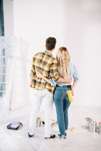 vista trasera de la joven pareja abrazando mientras mira a la pared blanca en casa
 - Foto, Imagen