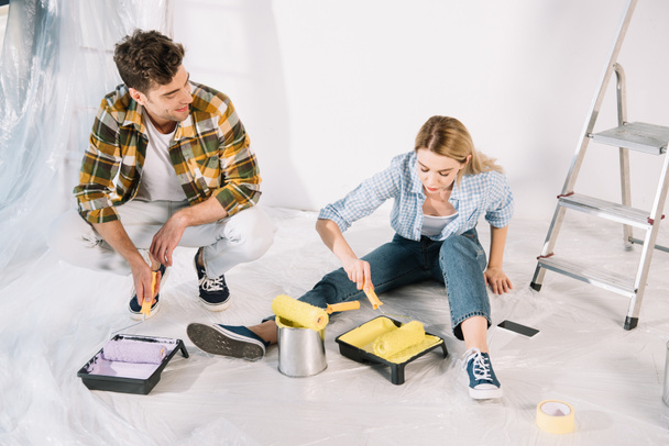 handsome man holding pink paint roller and woman putting paint roller into yellow paint while sitting on floor - Photo, Image