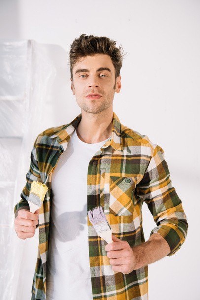 handsome young man looking at camera while holding yellow and pink paintbrushes - Фото, изображение