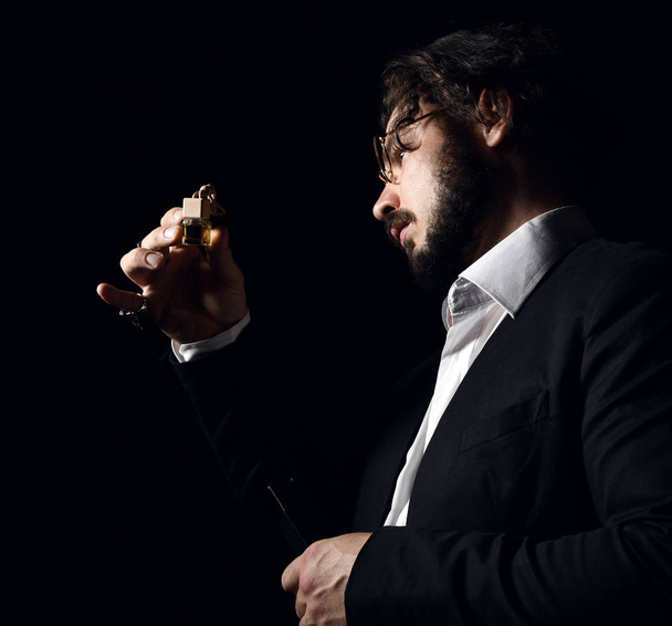 Brutal man in glasses with beard and modern hairstyle perfumer examines small bottle of perfume fragrance on dark - Φωτογραφία, εικόνα
