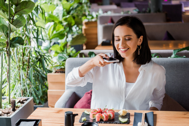 happy woman taking photo of tasty sushi in restaurant  - Fotografie, Obrázek