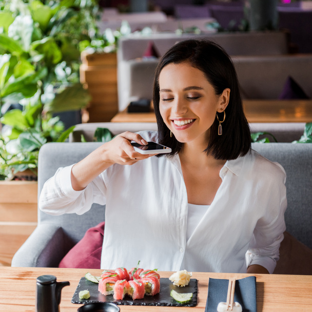 happy woman taking photo of delicious sushi in restaurant  - Фото, зображення