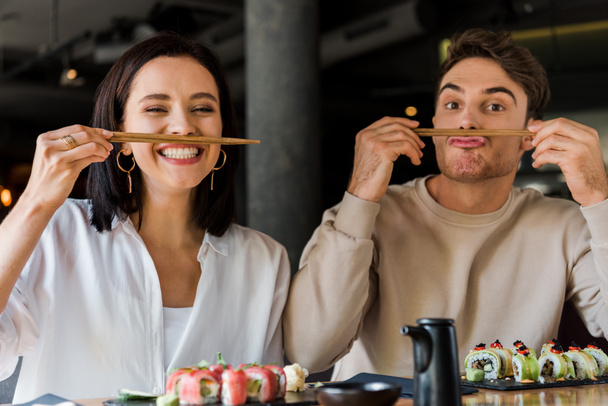schöner Mann mit Essstäbchen in der Nähe des Gesichts, während er mit fröhlichen Mädchen in der Sushi-Bar sitzt  - Foto, Bild