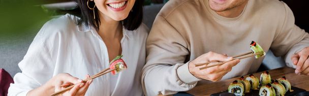 panoramiczne ujęcie szczęśliwego mężczyzny i kobiety trzymających pałeczki ze smacznym sushi w restauracji  - Zdjęcie, obraz