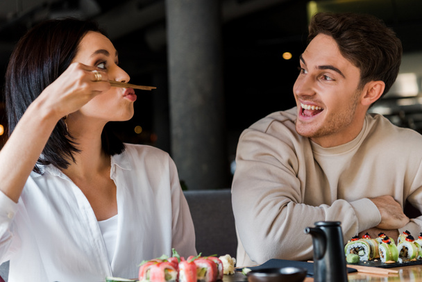 messa a fuoco selettiva della donna che tiene le bacchette vicino al viso mentre siede con l'uomo allegro nel sushi bar
  - Foto, immagini