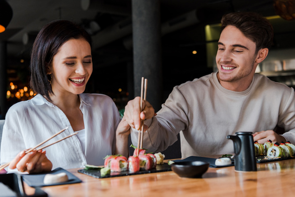 Selektiver Fokus einer glücklichen Frau mit Essstäbchen und leckerem Sushi in der Nähe eines gutaussehenden Mannes im Restaurant  - Foto, Bild