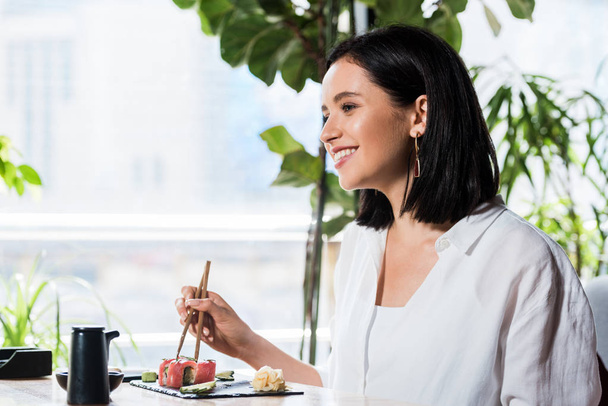 young happy woman holding chopsticks near delicious sushi in restaurant  - Photo, Image