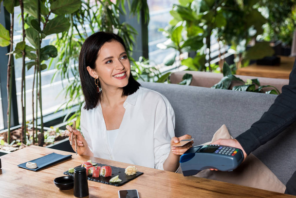 przycięty widok kelnera trzymającego czytnik kart kredytowych w pobliżu atrakcyjnej kobiety płacącej kartą kredytową  - Zdjęcie, obraz