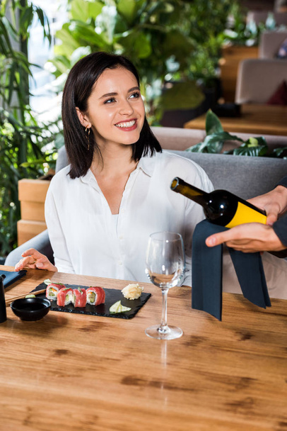 vista recortada de camarero sosteniendo botella con vino cerca de vidrio y mujer feliz en sushi bar
  - Foto, Imagen