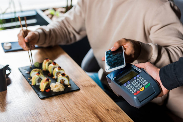 περικοπή άποψη της σερβιτόρας κρατώντας τερματικό πληρωμής κοντά στον άνθρωπο με smartphone σε σούσι μπαρ  - Φωτογραφία, εικόνα