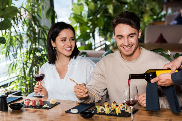 oříznutý pohled číšníka nalévajícího červené víno ve skle poblíž muže a ženy v sushi baru  - Fotografie, Obrázek