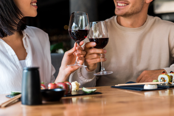 vista recortada de hombre feliz y mujer alegre tintineo vasos con vino tinto cerca de sushi
  - Foto, Imagen