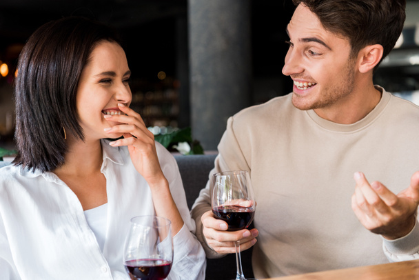 happy man looking at woman laughing while holding glasses with red wine  - Foto, imagen