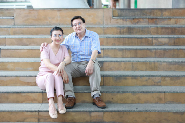 アジアの旅行者夫婦は、ダウンタウンの階段に座って白い素敵な帽子をかぶったピンクと青のシャツを着て、抱きしめながら幸せな笑顔で休憩します  - 写真・画像