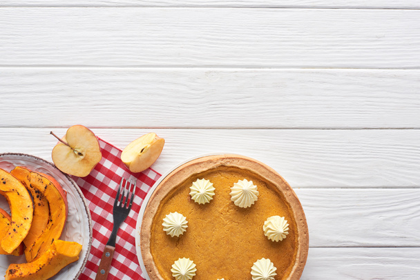 tarte à la citrouille savoureuse avec crème fouettée sur une serviette à carreaux près de tranches de citrouille cuite au four, pomme coupée et fourchette sur une table en bois blanc
 - Photo, image