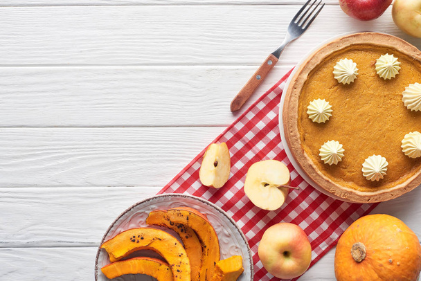 tasty pumpkin pie with whipped cream on checkered napkin near raw and sliced baked pumpkins, cut and whole apples, and fork on white wooden table - Photo, Image