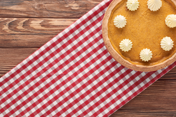 top view of delicious pumpkin pie with whipped cream on checkered napkin on brown wooden surface - Foto, imagen