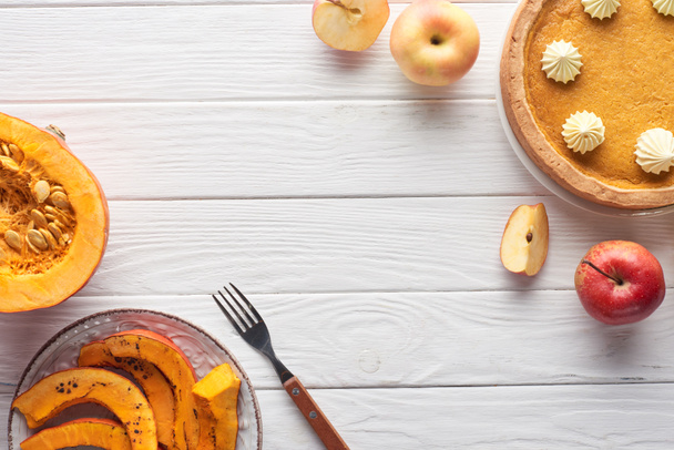 вкусный тыквенный пирог со взбитыми сливками рядом с сырой и нарезанной запечённой тыквой, яблоками и вилкой на белом деревянном столе
 - Фото, изображение