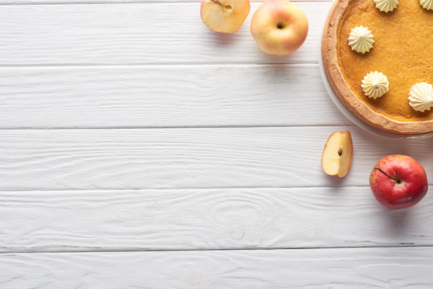 top view of delicious pumpkin pie with whipped cream near cut and whole apples on white wooden table - Fotoğraf, Görsel