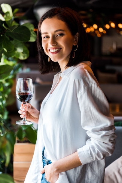 cheerful woman standing with hand in pocket while holding glass with red wine  - Fotó, kép