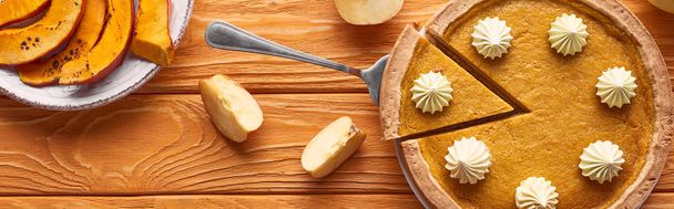 panoramiczny strzał smacznego ciasta dyniowego z bitą śmietaną w pobliżu pokrojonej w plasterki pieczonej dyni i pokrojone jabłko na pomarańczowym drewnianym stole - Zdjęcie, obraz