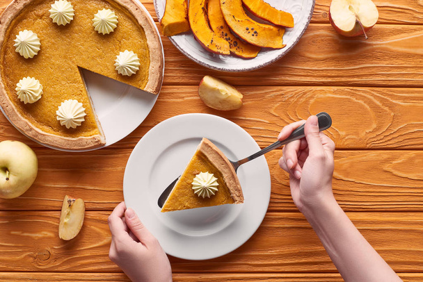 cropped view of woman holding spatula with piece of pumpkin pie with whipped cream near baked pumpkin, cut and whole apples on orange wooden table - Photo, image