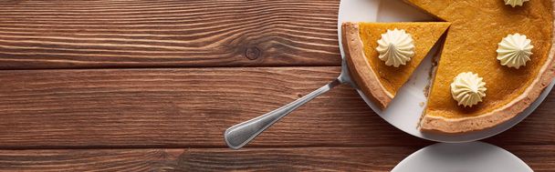 panoramic shot of delicious pumpkin pie with whipped cream on plate with spatula on brown wooden table - Photo, Image