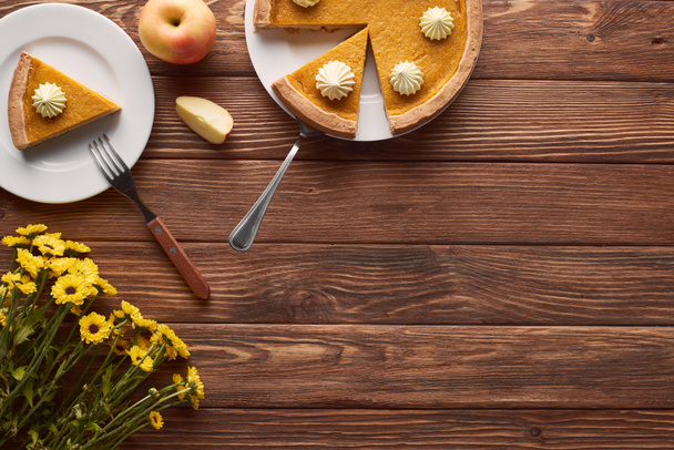 délicieuse tarte à la citrouille avec crème fouettée sur des assiettes avec spatule et fourchette près de pommes coupées et entières, et fleurs jaunes sur table en bois marron
 - Photo, image