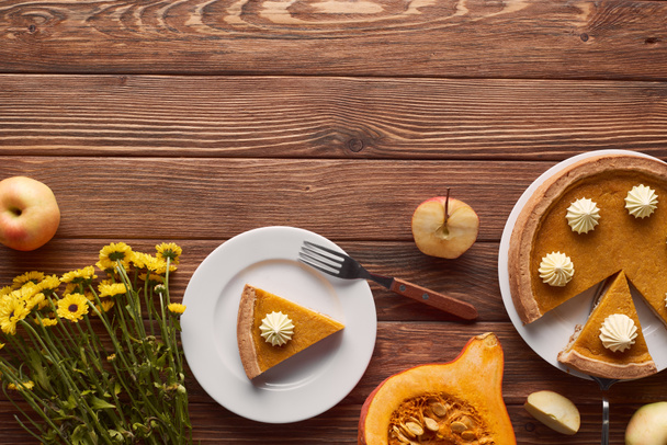 tasty pumpkin pie with whipped cream near fork, half of raw pumpkin, cut and whole apples, and yellow flowers on brown wooden table - Foto, immagini